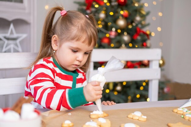 linda niña en pijama cocinando pan de jengibre festivo en la cocina decorada con navidad