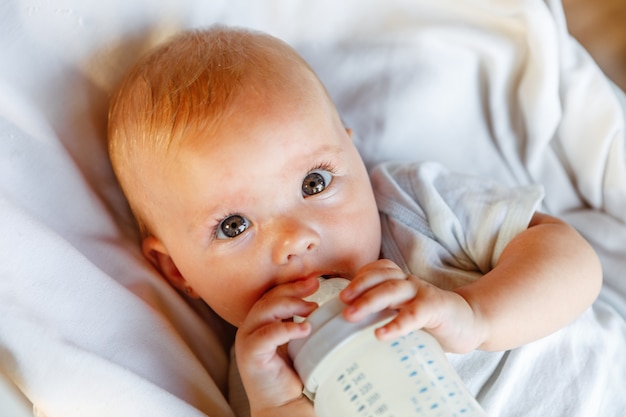 Linda niña recién nacida bebiendo leche de botella y mirando a cámara sobre fondo blanco. Bebé bebé chupando comer leche nutrición acostado en la cama de la cuna en casa. Concepto de niño feliz de maternidad.