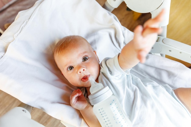 Linda niña recién nacida bebiendo leche de la botella y mirando a la cámara sobre fondo blanco Bebé infantil chupando comer leche nutrición acostado en la cama de la cuna en casa Concepto de niño feliz de maternidad