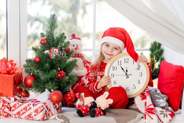 Una linda niña con un reloj en sus manos está sentada en el alféizar de la ventana de la casa junto al árbol de Navidad y esperando el año nuevo o la Navidad con un sombrero de Santa Claus y sonriendo