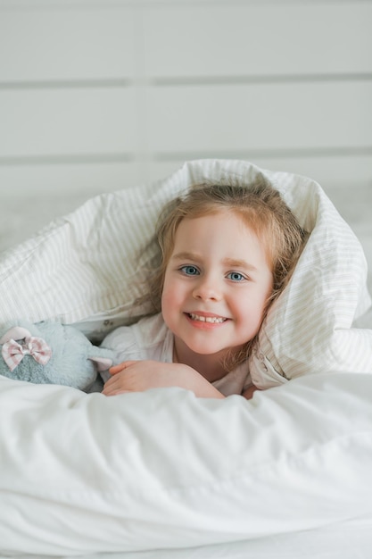 Una linda niña rizada de ojos azules juega en la cama con su peluche El niño está acostado en la cama debajo de las sábanas Emociones Sueño saludable