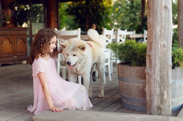 Linda niña rizada en vestido rosa con un gran perro blanco sentado en el balcón de la terraza abraza a su perro favorito durante una caminata de verano Concepto de estilo de vida