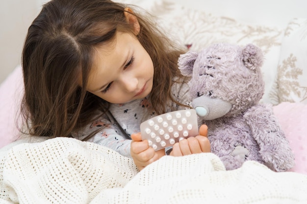 Linda niña en ropa de dormir sentada debajo de una manta en el sofá y tratando a un oso de peluche con pastillas