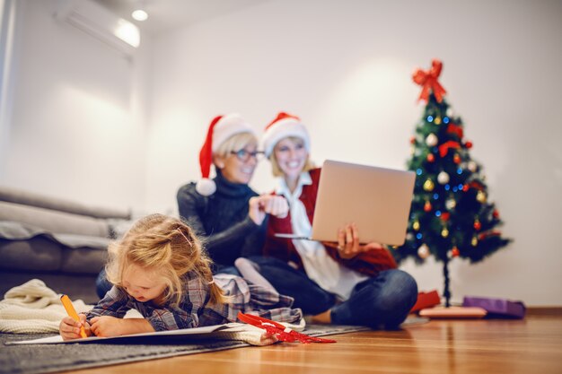 Linda niña rubia acostada sobre el estómago en el piso y el dibujo. En el fondo están la abuela y la madre usando una computadora portátil. La Navidad es tiempo para la familia.