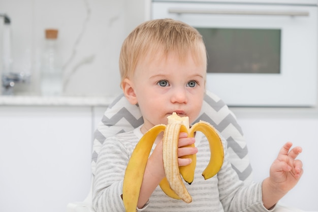 Linda niña rubia caucásica de un año de edad sentada en una silla almorzando en la cocina ...