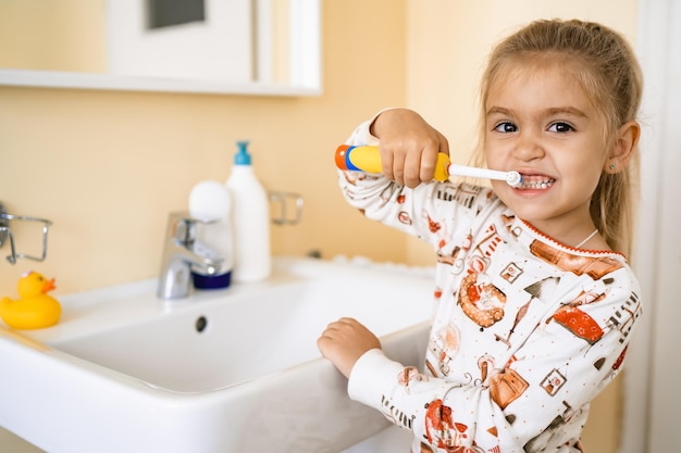 Foto una linda niña rubia se está cepillando los dientes con un cepillo eléctrico en el baño