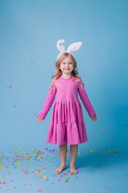 Linda niña rubia en orejas de conejo de Pascua en vestido rosa sobre fondo azul.