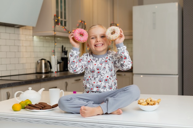 Linda niña rubia en ropa estampada de Navidad está sentada en la hermosa cocina con dos donas