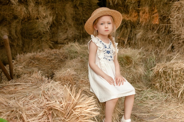 Linda niña rubia con vestido ligero con bordado y sombrero de paja sentado en un pajar en un pajar en un día soleado de verano Concepto de vacaciones rurales