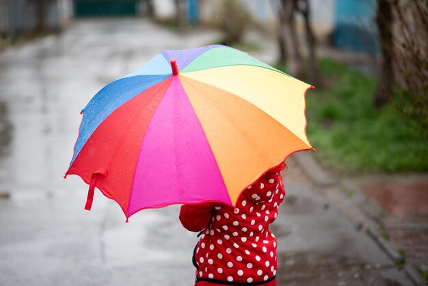 Una linda niña salta en charcos y se divierte La niña tiene un paraguas de arcoíris en sus manos