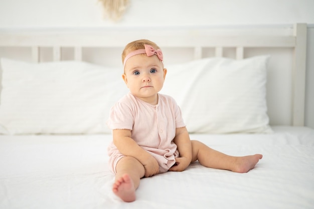 Una linda niña sana de hasta un año de edad con un traje rosa hecho de tela natural está sentada en una cama con ropa de cama blanca en el dormitorio mirando a la cámara, el bebé está en casa