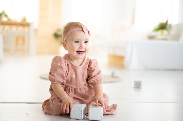 Una linda niña sana de hasta un año con un vestido rosa hecho de tela natural está sentada en una alfombra en la sala de estar de la casa jugando con cubos de madera el desarrollo del niño en casa