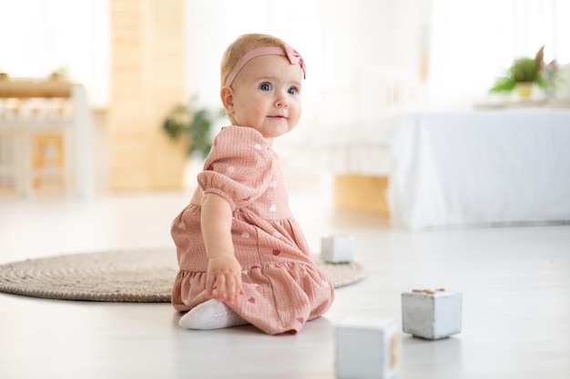 Una linda niña sana de hasta un año con un vestido rosa hecho de tela natural está sentada en una alfombra en la sala de estar de la casa jugando con cubos de madera el desarrollo del niño en casa