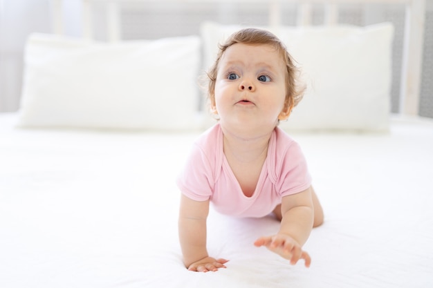 Linda niña sana en un traje rosa se arrastra sobre la cama, sobre ropa de cama blanca, sonriendo