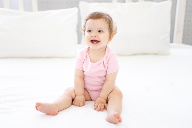 Una linda niña sana con un traje rosa está sentada en la cama con ropa de cama blanca sonriendo