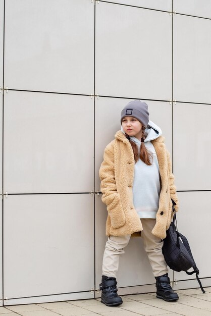 una linda niña de seis años con un abrigo de piel sintética beige está parada contra una pared en la ciudad