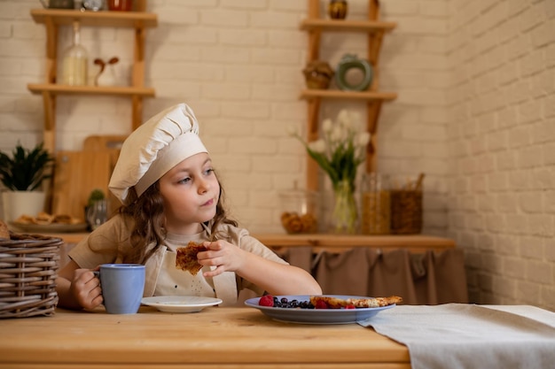 una linda niña de seis años con sombrero y delantal de chef está sentada en una mesa de madera comiendo panqueques