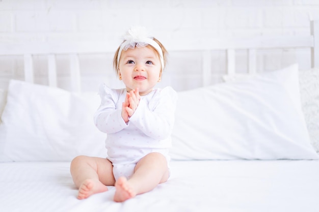 Linda niña sentada en la cama con ropa blanca y con un lazo en la cabeza y aplaudiendo o bailando un bebé divertido en una cama de algodón en casa y sonriendo el concepto de productos para bebés