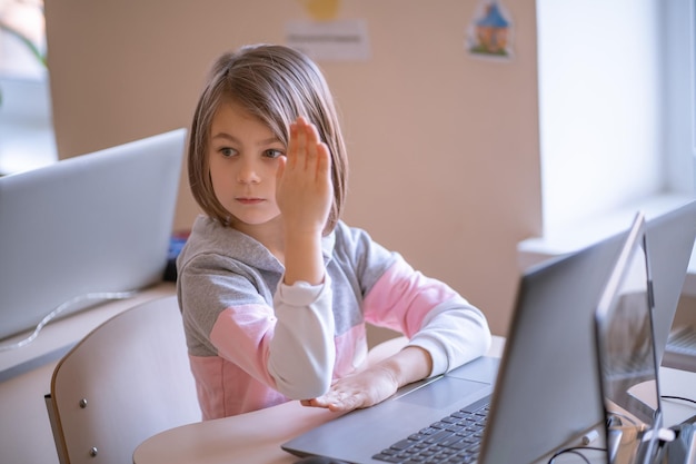 Linda niña sentada en el escritorio de la escuela con una computadora portátil levanta la mano para responder