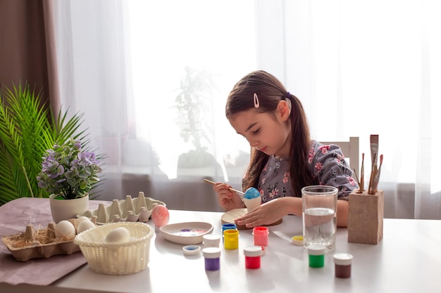 Una linda niña sentada en una mesa blanca junto a la ventana pinta huevos con un pincel y pinta en frascos