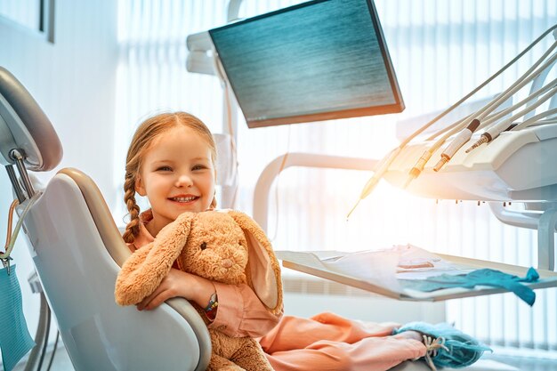 Una linda niña sentada en un sillón dental sosteniendo un conejo de juguete y riendo En el fondo equipo dental y un monitor para el logotipo de la clínica Medicina para el cuidado dental Luz del sol