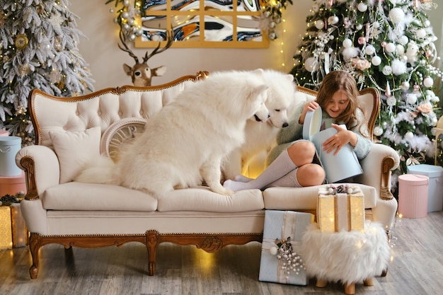 Linda niña sentada en el sofá con dos perros Samoyedo, abriendo cajas de regalo cerca del árbol de Navidad en un hermoso interior