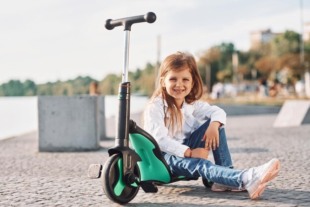 Linda niña sentada y sonriente con scooter está al aire libre en verano