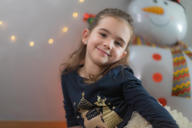 Linda niña sentada en el suelo con expresión amable globo de Navidad y decoración de luces en el fondo
