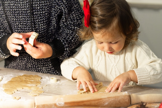 Linda niña seria 2-4 con un lazo rojo prepara galletas de jengibre de Navidad en la cocina de Año Nuevo.