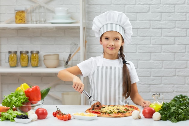Linda niña con sombrero de chef y un delantal cortando pizza