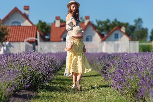 Una linda niña con sombrero de paja y vestido amarillo corre hacia su hermosa madre en vestido de verano en casa