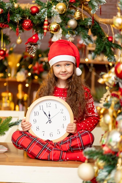 Una linda niña con un sombrero de Papá Noel y un suéter rojo con un reloj en sus manos se ríe de felicidad en la oscura cocina de la casa en previsión del año nuevo y la Navidad