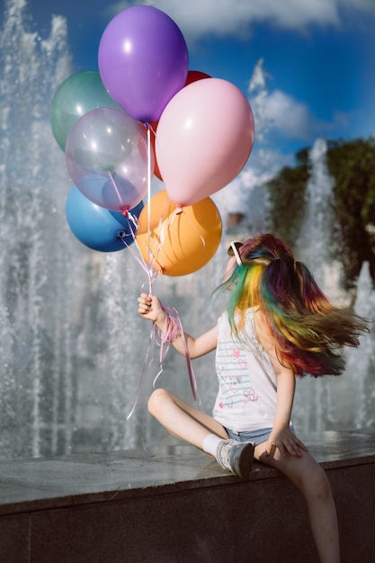 Linda niña sonriente caucásica con el pelo teñido de colores vistiendo gafas de sol mamás sosteniendo globos sentado