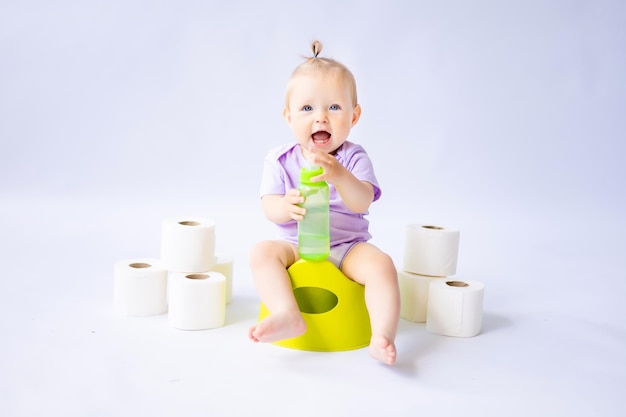 Una linda niña sonriente está sentada en una olla con una botella de agua y rollos de papel higiénico aislados en fondo blanco