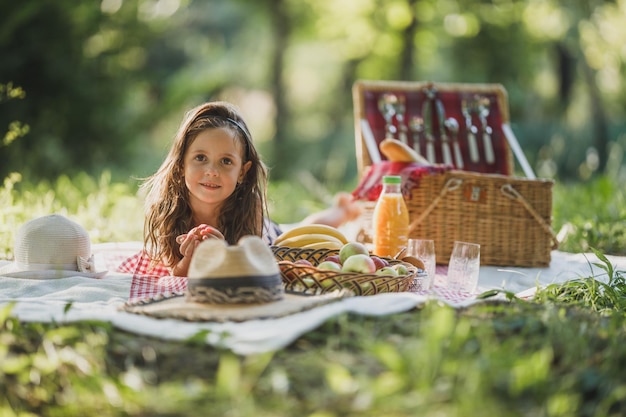 Una linda niña sonriente está tumbada en la manta sobre un césped y disfruta del día de picnic en el parque.