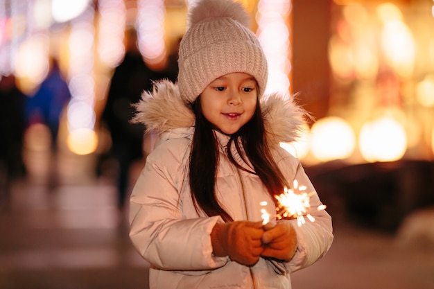 Linda niña sonriente con luces de bengala en la calle por la noche en invierno