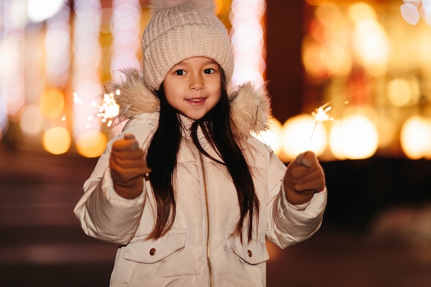 Linda niña sonriente con luces de bengala en la calle por la noche en invierno