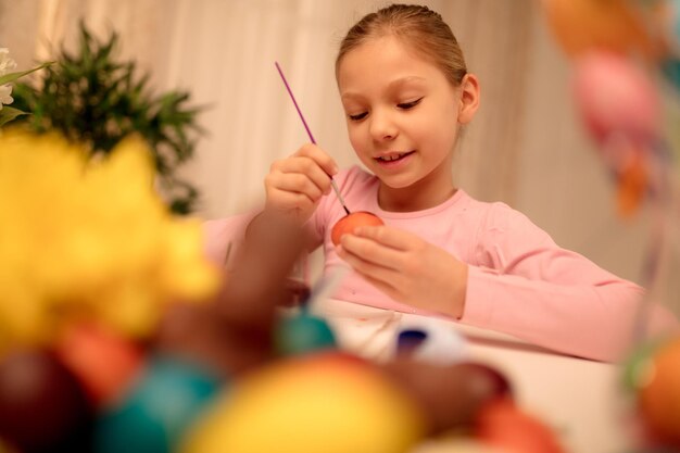 Linda niña sonriente pintando huevos de Pascua en casa.