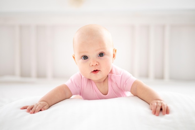 Una linda niña sonriente con un traje rosa está acostada en la cama en el dormitorio de la casa con ropa de cama blanca boca abajo mirando a la cámara sonriendo feliz bebé