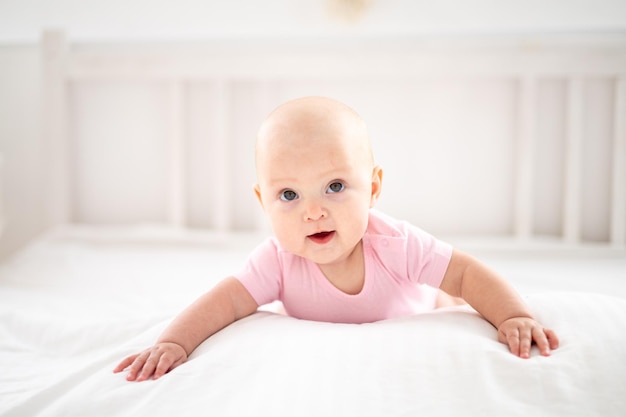 Una linda niña sonriente con un traje rosa está acostada en la cama en el dormitorio de la casa con ropa de cama blanca boca abajo mirando a la cámara sonriendo feliz bebé