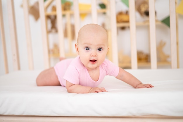 Una linda niña sonriente con un traje rosa está acostada en una cuna en casa sobre ropa de cama blanca boca abajo mirando a la cámara sonriendo feliz bebé