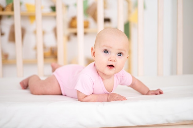 Una linda niña sonriente con un traje rosa está acostada en una cuna en casa sobre ropa de cama blanca boca abajo mirando a la cámara sonriendo feliz bebé