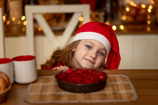 Una linda niña con un suéter rojo y un gorro de Papá Noel está en la cocina bebiendo té con un pastel y esperando el año nuevo o la Navidad