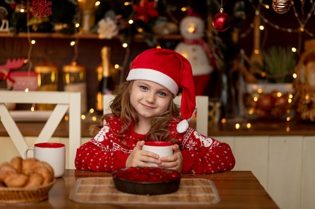 Una linda niña con un suéter rojo y un gorro de Papá Noel está en la cocina bebiendo té con un pastel y esperando el año nuevo o la Navidad