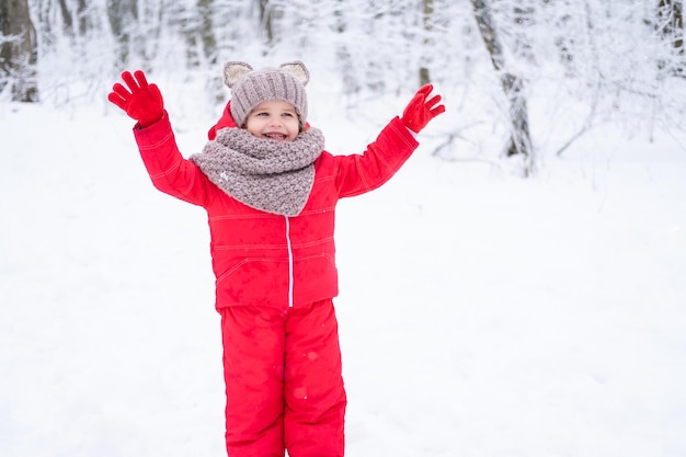 Linda niña en traje de nieve rosa y gorro de punto y bufanda juega con nieve en el bosque de invierno