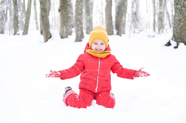 Linda niña en traje de nieve rosa y gorro de punto y bufanda juega con nieve en el bosque de invierno