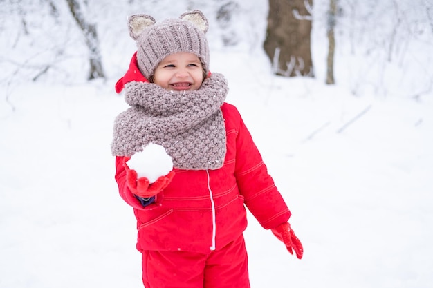 Linda niña en traje de nieve rosa y gorro de punto y bufanda juega con nieve en el bosque de invierno