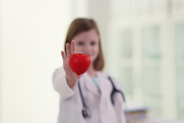 Linda niña vestida como médico tiene un juguete de corazón rojo en la mano, un niño con estetoscopio se para en