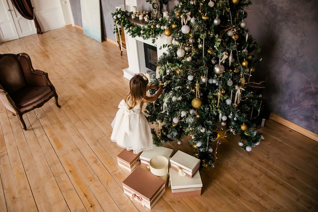 Linda niña en el vestido blanco con grandes regalos cerca del árbol de Navidad