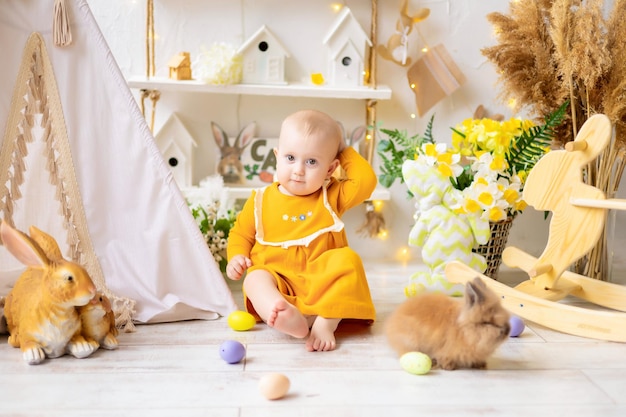 Una linda niña con un vestido naranja está sentada en casa con huevos de Pascua entre flores con conejos y accesorios de Pascua Celebración de la Gran Pascua Tradiciones de Pascua